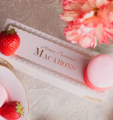 A pink and red box with a clear lid displays six delicate macarons in shades of pink and white. The macarons are perfectly round with a delicate, crisp exterior and a soft, chewy interior. The filling of the macarons is visible through the clear lid, revealing a creamy, pink filling that is flecked with red. A pink ribbon tied around the box adds a touch of elegance and makes it the perfect gift for Valentine's Day. aimee provence